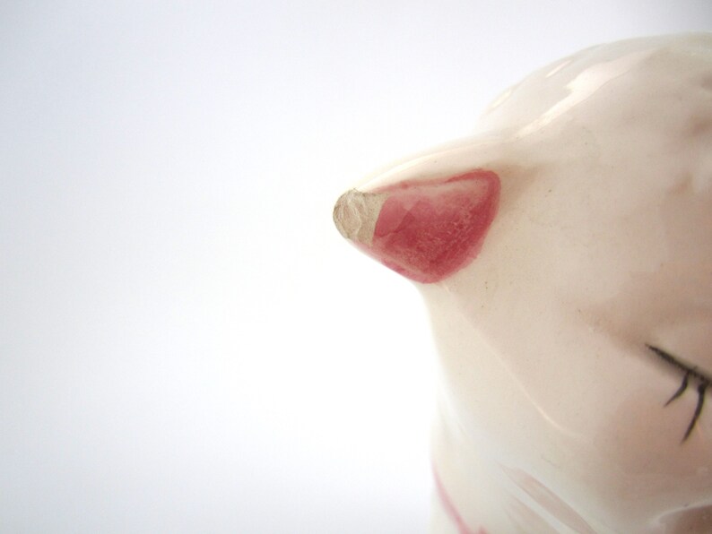 May include: Close-up of a white ceramic piggy bank with a pink ear and black painted details. The piggy bank is slightly chipped on the ear.
