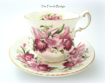 Puede incluir: Una taza de té y un platillo blancos con un patrón floral rosa y morado. El diseño floral presenta orquídeas con hojas verdes. La taza y el platillo tienen un borde dorado.