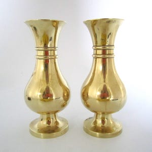 May include: Two brass vases with a shiny, reflective surface. Each vase has a flared top, a bulbous midsection, and a round base. The vases are a warm gold color and appear to be vintage or antique.