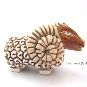 Può includere: Una scultura in ceramica di una pecora bianca con corna marroni. La pecora ha un corpo dettagliato e testurizzato e un viso stilizzato.