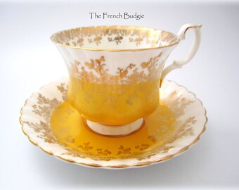 May include: A yellow teacup and saucer with gold trim and floral pattern. The teacup has a handle and the saucer has a scalloped edge.
