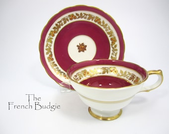 Può includere: Un set vintage di tazza da tè e piattino di colore rosso cremisi intenso con finiture dorate. Il set presenta un delicato motivo floreale con foglie dorate e bacche arancioni.