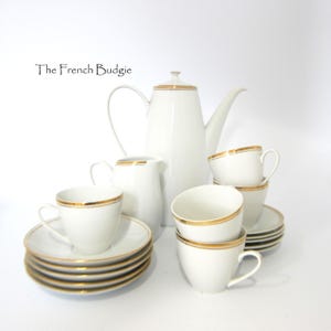 May include: A white porcelain tea set with gold trim. The set includes a tall coffee pot, creamer, sugar bowl, saucers, and several teacups. The text "The French Budgie" is in the upper left corner.