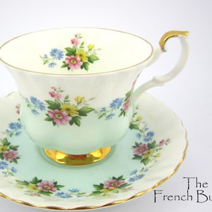 May include: A vintage teacup and saucer set. The cup and saucer are decorated with floral patterns in pink, yellow, and blue on a light green and white background. The cup has a gold handle and a gold rim.