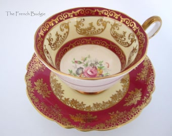 Pode incluir: Um conjunto de chá vintage de xícara e pires com um design floral vermelho e dourado. A xícara tem uma alça delicada e uma borda festonada. O pires tem um design correspondente e uma borda festonada.