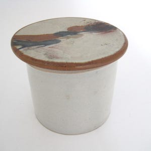 Peut inclure: Petit pot en céramique blanc cassé avec un couvercle rond. Le couvercle présente un bord marron et un motif peint avec des formes abstraites marron, noires et rouges. Le pot est cylindrique et de conception minimaliste.