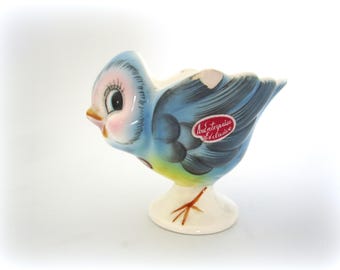 Op de afbeelding: Een vintage keramieken vogelbeeldje in tinten blauw, roze en geel. De vogel heeft grote ogen, een snavel en een kleine rode sticker met de tekst "An Enterprise Exclusive". Het beeldje staat op een witte voet met bruine poten.