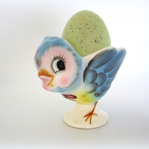 May include: A ceramic egg cup in the shape of a bluebird, holding a speckled green egg. The bird features a pink face, blue wings, and a yellow belly. The egg cup is on a white pedestal base.
