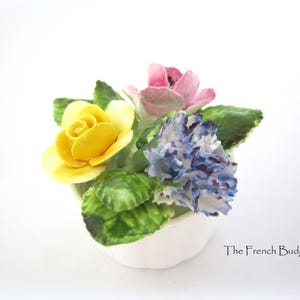 Może przedstawiać: Mały biały ceramiczny doniczkowy z żółtą różą, różowym kwiatem i niebiesko-białym goździkiem.