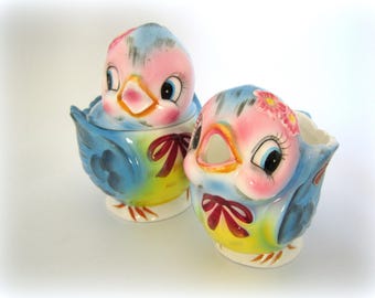 May include: A blue and yellow bird-shaped sugar bowl and creamer set with pink and white accents. The birds have large eyes and a red bow.