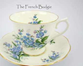 Könnte beinhalten: Eine weiße Teetasse und Untertasse mit einem floralen Muster aus blauen Vergissmeinnicht-Blüten und grünen Blättern. Tasse und Untertasse haben einen goldenen Rand.