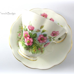 Peut inclure: Une tasse à thé et une soucoupe blanches avec un motif floral composé de fleurs roses et de feuilles vertes. La tasse et la soucoupe ont une bordure dorée.