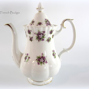 May include: White porcelain teapot with a floral pattern of purple violets. The teapot has a gold trim around the lid and base.