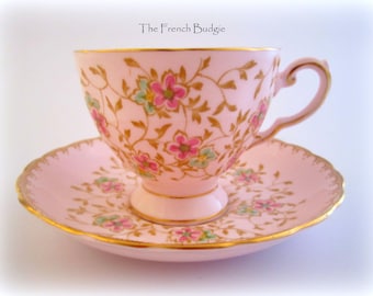 May include: A pink teacup and saucer set with a delicate floral pattern in shades of pink, green, and gold. The set features a gold rim and a scalloped edge on the saucer.
