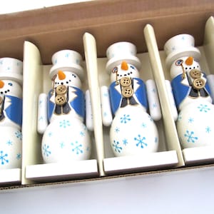 Op de afbeelding: Een set houten sneeuwpopfiguren in een kartonnen doos. Elke sneeuwpop heeft een witte body met blauwe sneeuwvlokaccenten, een blauwe jas en een witte hoge hoed. Ze hebben oranje wortelneuzen en gouden knoopdetails.