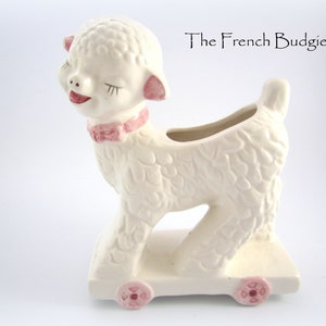 May include: White ceramic lamb planter with pink details, on wheels, with a smiling face and closed eyes. The lamb is on a white base with pink wheels. The text "The French Budgie" is visible in the background.