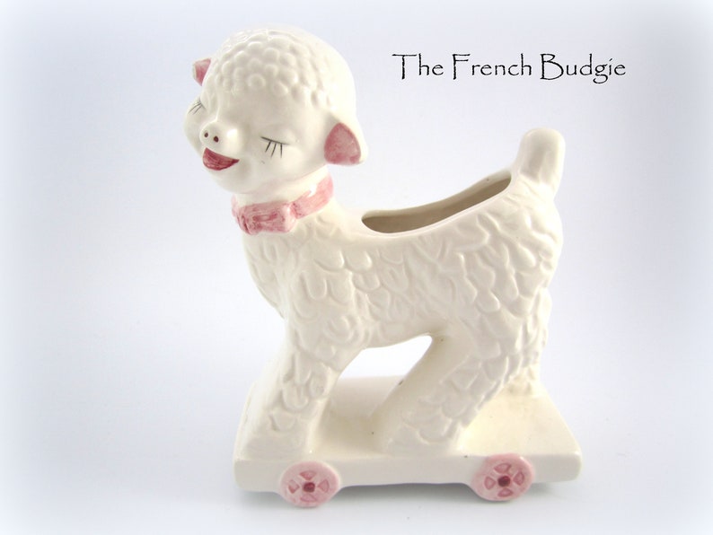 May include: White ceramic lamb planter with pink details, on wheels, with a smiling face and closed eyes. The lamb is on a white base with pink wheels. The text "The French Budgie" is visible in the background.