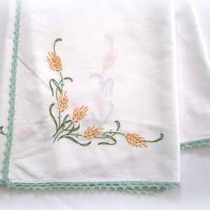 Könnte beinhalten: Weiße Leinen-Tischdecke mit grünem Häkelrand und gesticktem Blumendesign mit orangefarbenen Weizenähren.