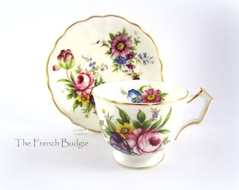 Puede incluir: Una taza de té y un platillo blancos con un patrón floral. La taza y el platillo tienen un borde dorado. Las flores son rosas, amarillas, azules y moradas.
