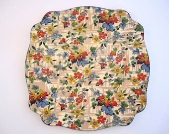 May include: A vintage floral patterned cake plate with a scalloped edge. The plate features a white background with a repeating pattern of red, yellow, blue, and green flowers and leaves.
