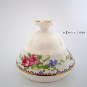 May include: A white ceramic trinket box lid with a floral cross-stitch design in pink, blue, and green. The lid has a gold rim and a decorative flower-shaped handle. The text "The French Budgie" is visible.