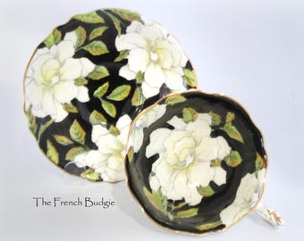 May include: A teacup and saucer set featuring a black background adorned with a white floral pattern. The flowers are intricately designed, complemented by green leaves. The cup and saucer are edged with gold.