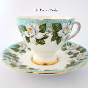 May include: A vintage teacup and saucer set. The cup and saucer are white with a turquoise rim and floral design. The teacup features a gold-colored base and handle. The text "The French Budgie" is at the top.