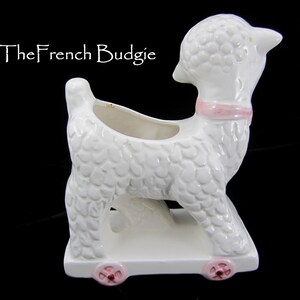 May include: White ceramic planter shaped like a lamb with pink wheels and a pink ribbon around its neck. The planter has a textured surface that resembles fur.