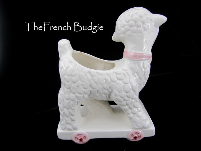 May include: White ceramic planter shaped like a lamb with pink wheels and a pink ribbon around its neck. The planter has a textured surface that resembles fur.
