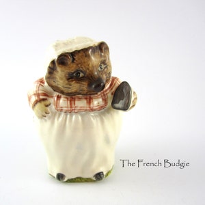 May include: A ceramic figurine of a hedgehog wearing a red and white checked dress and a white apron, holding a brown iron. The figurine is standing on a green base.