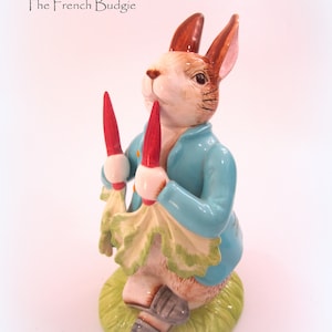Puede incluir: Figura de cerámica de un conejo con una chaqueta azul y dos zanahorias rojas. El conejo está sobre una base verde. El texto "The French Budgie" está en la esquina superior izquierda.