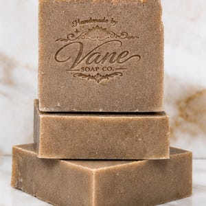May include: A stack of three rectangular bars of light brown soap. The top bar features a stamped logo that reads Handmade by Vane SOAP CO. with decorative flourishes and a lotus flower icon centered above the brand name in a dark brown ink.