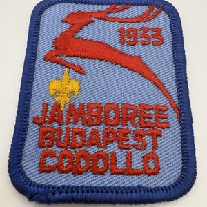 May include: A rectangular embroidered patch with a light blue background and a dark blue border. It features a red leaping deer, a yellow fleur-de-lis, and red text reading 1933 JAMBOREE BUDAPEST GODOLLO. The design is centered on the fabric patch.