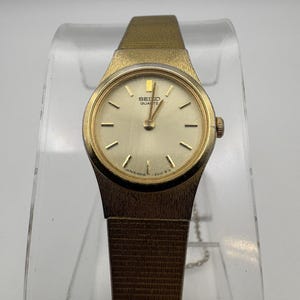 May include: A gold-tone Seiko Quartz wristwatch featuring a round champagne-colored dial with gold-tone hour markers and hands. The watch has a textured gold-tone metal case and a matching integrated mesh-style bracelet with a secure clasp.