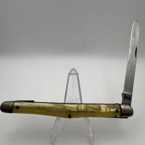 May include: A vintage folding pocket knife with a single silver-toned steel blade extended upward. The handle is made of a translucent yellow material with black text printed on it that reads L.R. ICE EXCHANGE PHONE 256 4551 COLUMBIA, S.C.