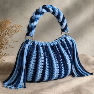 Puede incluir: Un bolso hecho de gruesas tiras de tela trenzada en tonos alternos de azul marino y azul claro. El bolso cuenta con un asa trenzada sujeta con anillos de metal plateado y largos flecos colgantes en ambos lados del cuerpo del bolso.