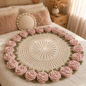May include: A circular crochet blanket featuring an intricate cream-colored center with a radiating openwork pattern. The border is adorned with a continuous ring of three-dimensional dusty pink crocheted roses nestled among small green leaves.