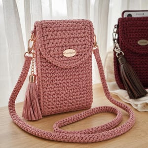 May include: A dusty rose crochet crossbody bag featuring a flap closure with a gold-tone oval tag labelled handmade. The bag includes a matching crochet strap and a decorative pink tassel attached to a gold-tone chain link on the side of the bag.