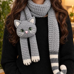 May include: A grey crocheted scarf featuring a cat head with green eyes, a pink nose, and white whiskers. The scarf has two front paws with white tips and a long, striped tail end with alternating grey and white bands, worn over a black jumper.