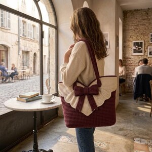 Op de afbeelding: Een diepbordeauxrode gehaakte tote bag met een grote, cr&egrave;mekleurige geschulpte flap en een opvallende bijpassende bordeauxrode strik in het midden. De tas heeft twee lange schouderbanden en een gestructureerd, geweven patroon over het geheel.