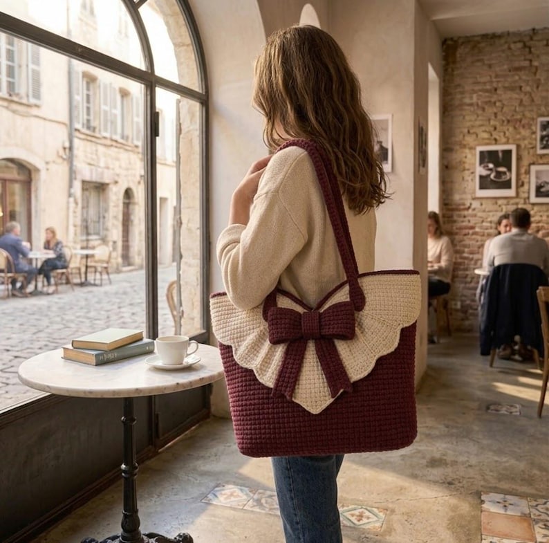 Op de afbeelding: Een diepbordeauxrode gehaakte tote bag met een grote, cr&egrave;mekleurige geschulpte flap en een opvallende bijpassende bordeauxrode strik in het midden. De tas heeft twee lange schouderbanden en een gestructureerd, geweven patroon over het geheel.