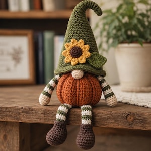 May include: A crochet gnome figure featuring a tall olive green pointed hat adorned with a yellow sunflower and a green leaf. The body is a round, ribbed orange pumpkin shape with a small beige nose. It has striped arms and legs in green and white.