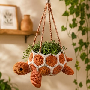 May include: A hanging crochet turtle planter featuring a shell constructed from orange hexagonal motifs outlined in white yarn. The turtle has a rounded head with a black eye, four flippers, and a tail, with three braided cords for suspension.