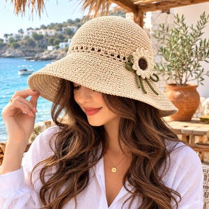 May include: A beige crochet sun hat featuring a wide brim and a decorative cream-colored flower applique with a dark brown center and green leaves attached to the side. The hat is constructed from a woven straw-like material with a textured pattern.