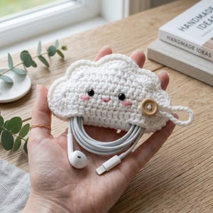 Puede incluir: Un organizador de cables de ganchillo blanco con forma de nube, con dos ojos de cuentas negras, mejillas bordadas en rosa y una pequeña boca rosa. Un botón de madera marrón claro asegura el lazo, con un cable de auriculares blanco enrollado.