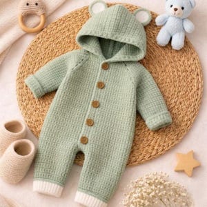 May include: A sage green knitted baby romper featuring a hood with bear ears, long sleeves, and a front button closure with five brown buttons. The garment has a textured waffle-knit pattern and white ribbed cuffs at the bottom of the legs.