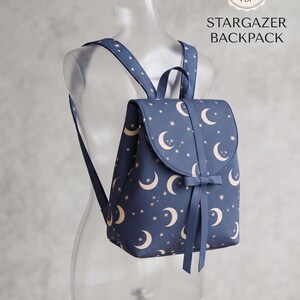 May include: A navy blue backpack featuring a repeating pattern of cream-coloured crescent moons and stars. The bag has a flap closure with a decorative bow and two shoulder straps. Text on the image reads: SEWING PATTERN PDF STARGAZER BACKPACK.