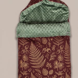 May include: A maroon baby sleeping bag featuring a gold-toned botanical print of ferns, acorns, and pinecones. The interior is lined with a sage green textured minky fabric. The hood includes two wooden toggles for closure and a fold-over design.