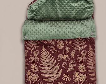 Op de afbeelding: Een kastanjebruine babyslaapzak met een goudkleurige botanische print van varens, eikels en dennenappels. De binnenzijde is gevoerd met een textuurrijke saliegroene minky-stof. De capuchon heeft twee houten knopen en een omgeslagen rand.