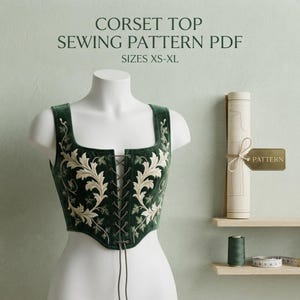 May include: A dark green corset top sewing pattern PDF in sizes XS-XL. The corset features a square neckline, a front lace-up closure with dark strings, and intricate cream-colored leaf embroidery. A rolled paper pattern and thread are visible.